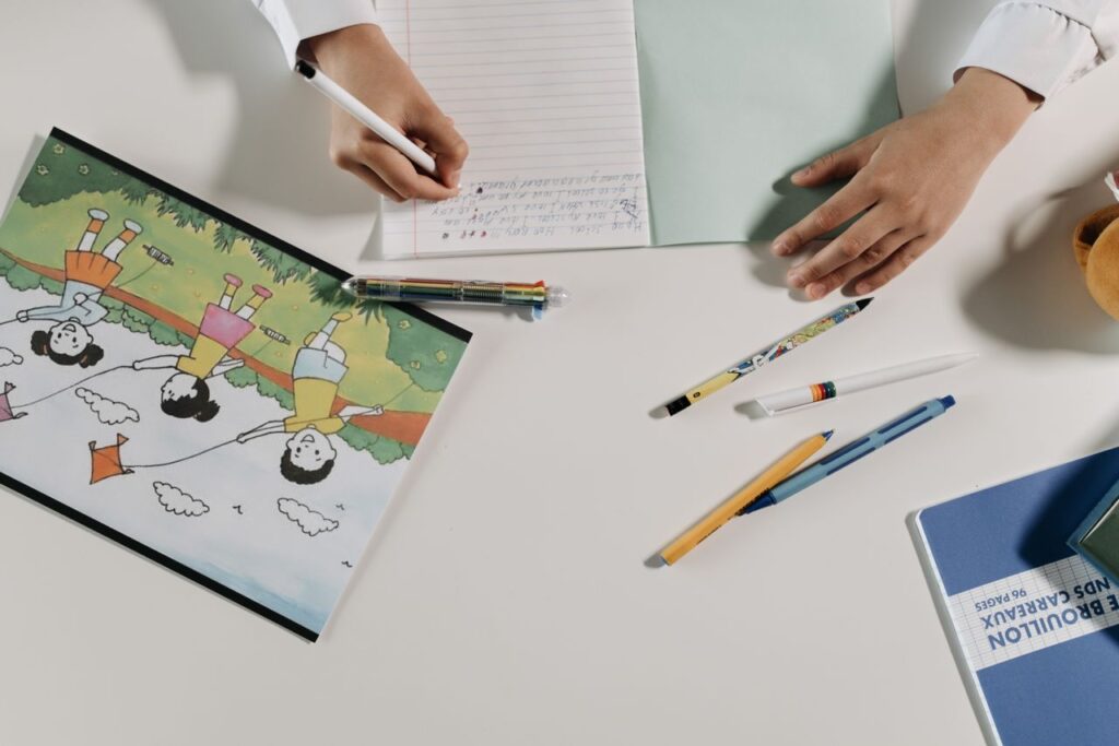 Child writing on notebook with stationery scattered on a desk, featuring colorful illustration.